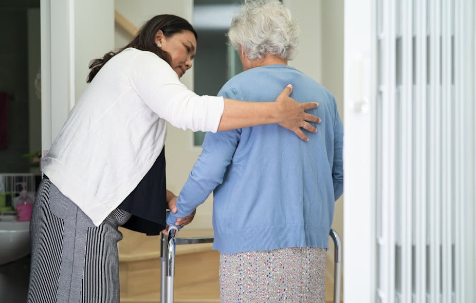 A caregiver helps support an elderly woman who is using a walker.