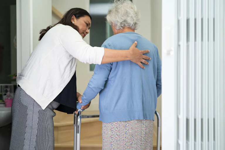 A caregiver helps support an elderly woman who is using a walker.