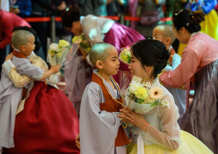 A young boy in a gray monk robe stands as a woman holding flowers kneels and smiles next to him.