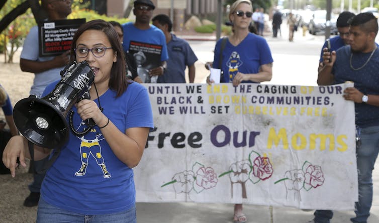 A woman speaks into a megaphone in front of a poster that says free our moms