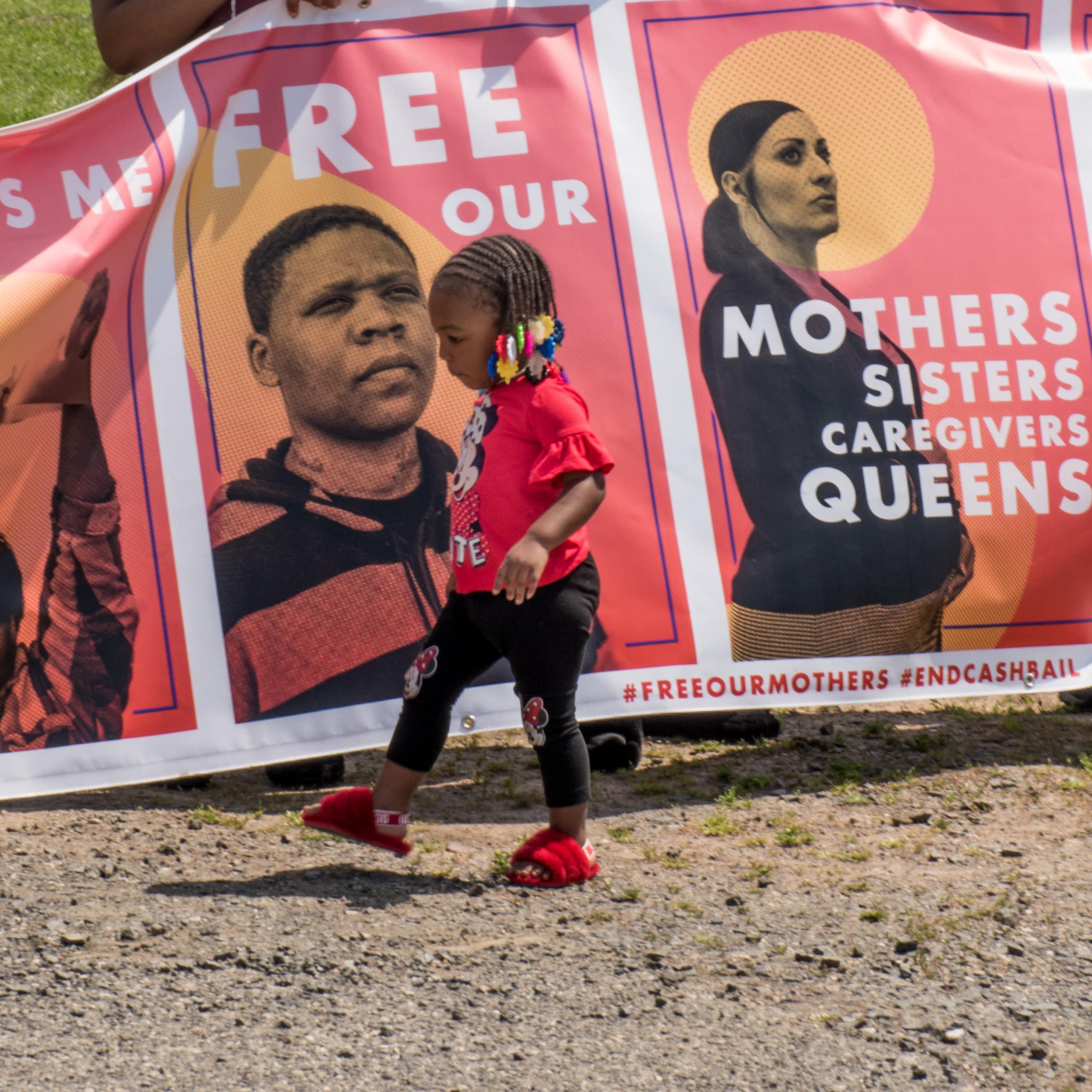 a toddler walks in front of a poster that says Free our Mothers