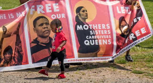 a toddler walks in front of a poster that says Free our Mothers