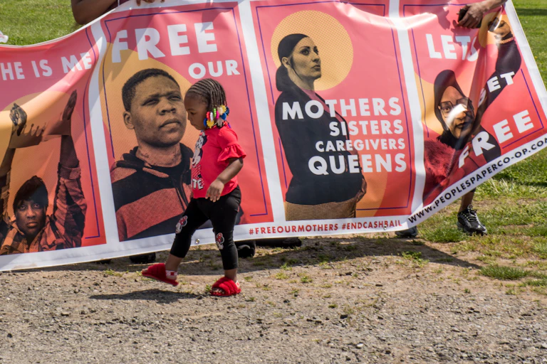 a toddler walks in front of a poster that says Free our Mothers