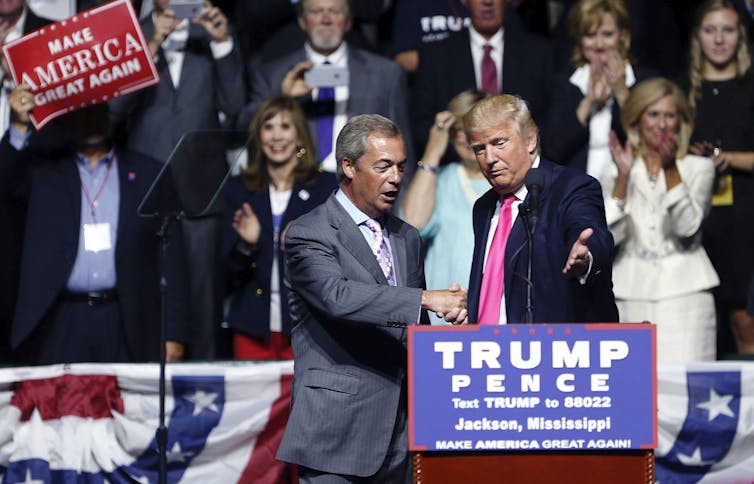 two men in suits shake hands on a stage. One invites the other to take the podium.