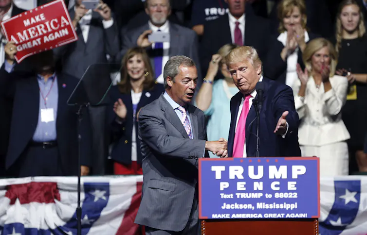 two men in suits shake hands on a stage. One invites the other to take the podium.
