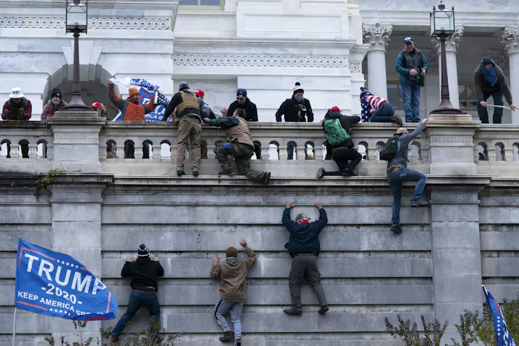People climb a wall. A Trump flag waves behind them.