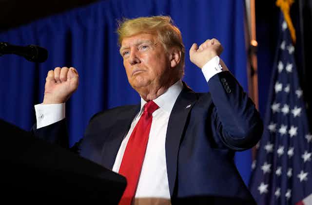 Donald trump wearing a dark suit and red tie raises his arms.