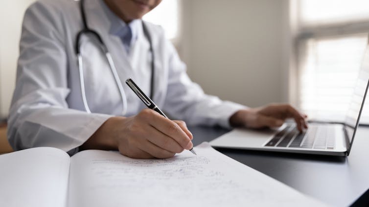 Doctor in white coat, stethoscope around neck, taking notes from laptop