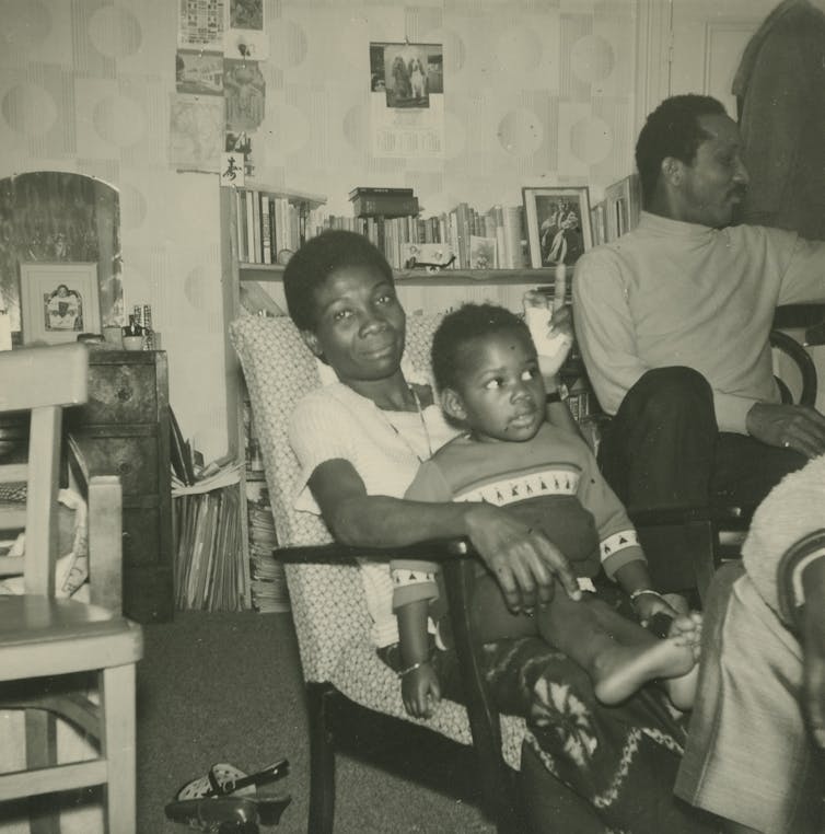 Jessica Huntley has daughter Accabre sat on her lap. Eric is to the right. They sit in a front room surrounded by books.