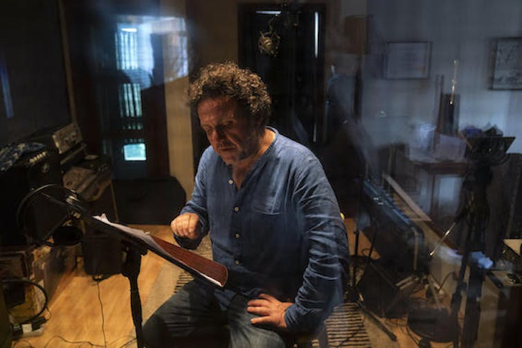 A man sits on a chair reading from papers on a music stand, music equipment all around him in a wooden-floored studio.