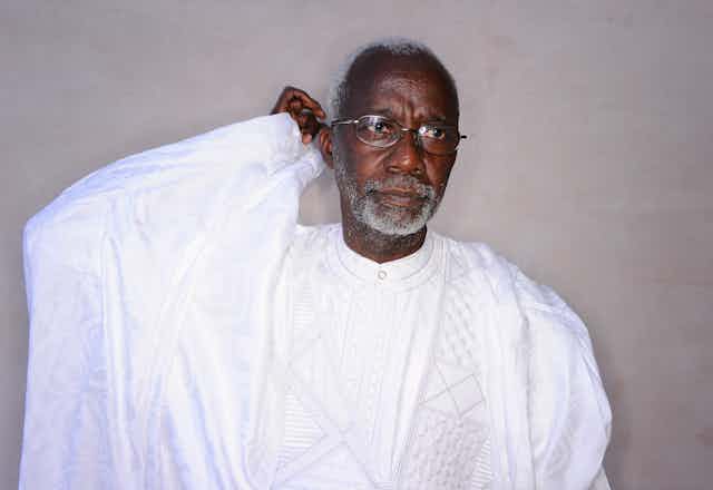 An elderly man with grey hair, beard and spectacles holds a hand behind his head as he looks into the distance. He is wearing a flowing white robe.