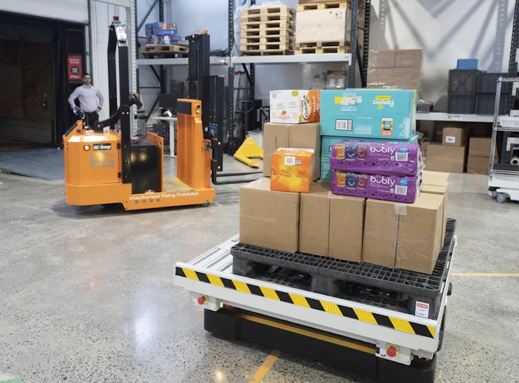 a photograph of a robot forklift and package mover which looks like a pallet on wheels with boxes stacked on top are seen on a factory floor