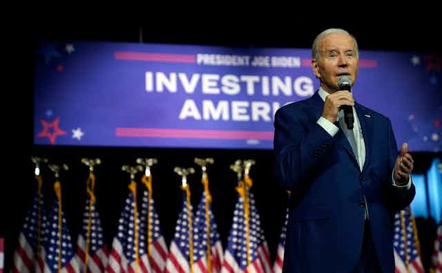 A grey-haired older man in a blue suit speaks into a microphone in front of a large banner that reads Investing in America.