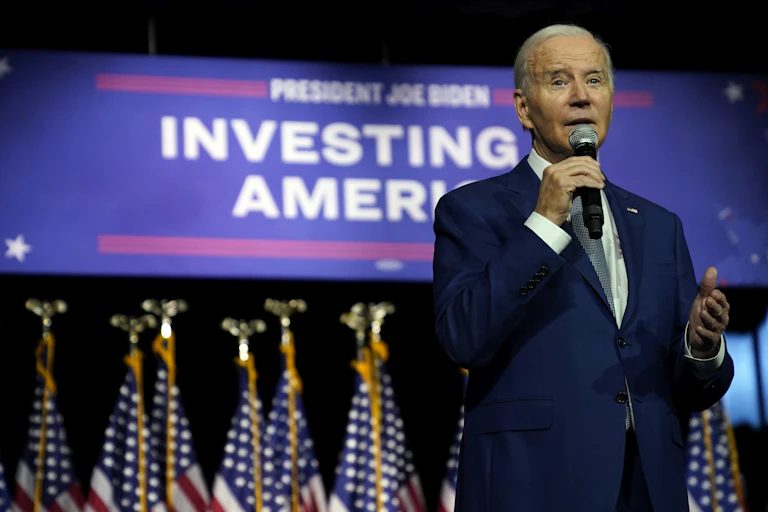 A grey-haired older man in a blue suit speaks into a microphone in front of a large banner that reads Investing in America.