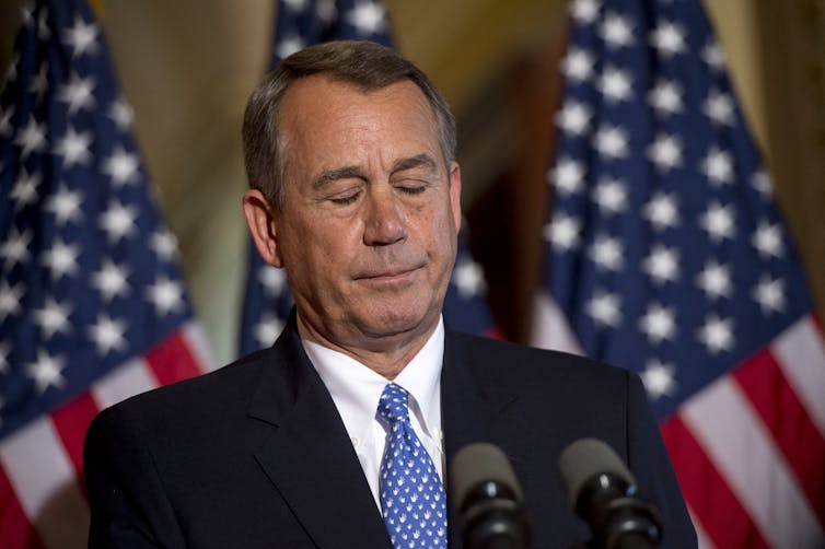 A middle-aged man in a suit, standing in front of several US flags with his eyes closed, looking glum.