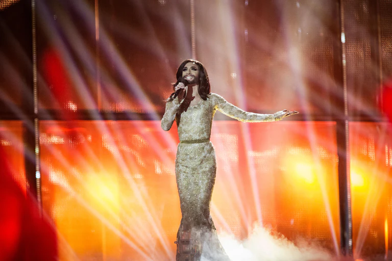 A transvestite in a beard and sparkly frock on stage singing.