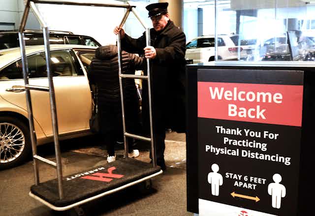 A man in a black hat and coat totes a Marriott hotel luggage cart next to a sign urging people to social distance and says 'welcome back.'