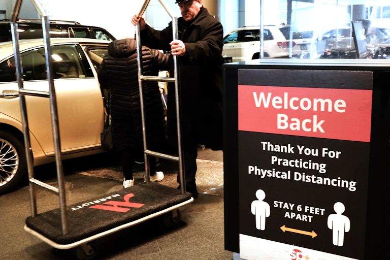 A man in a black hat and coat totes a Marriott hotel luggage cart next to a sign urging people to social distance and says 'welcome back.'