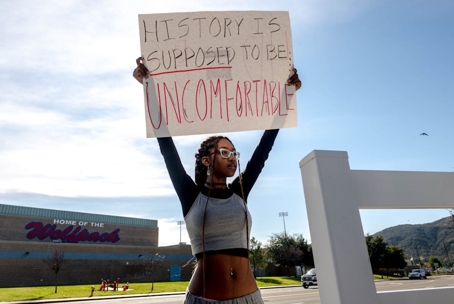 A Black teenage girl, wearing long braids, eyeglasses and a gray crop top, stands across the street from a high school. She holds a cardboard sign above her head that reads: History is supposed to be uncomfortable.