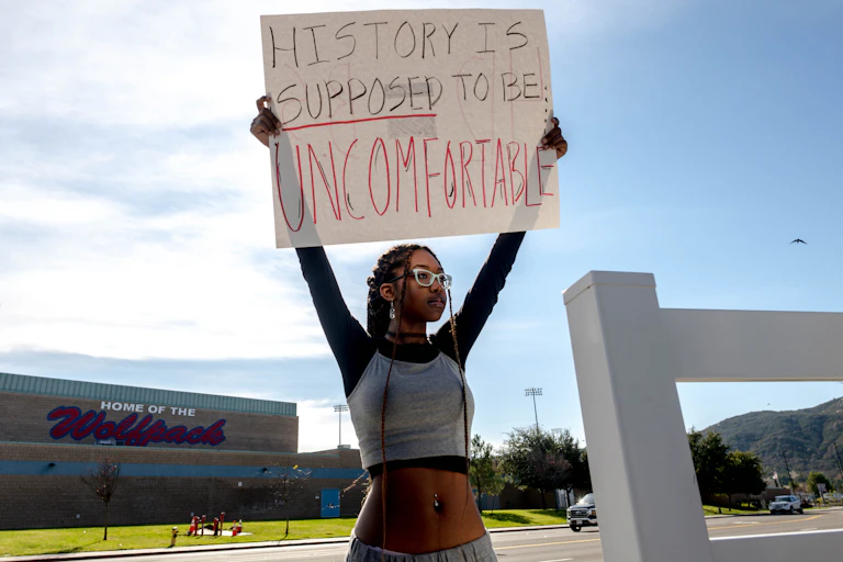 A Black teenage girl, wearing long braids, eyeglasses and a gray crop top, stands across the street from a high school. She holds a cardboard sign above her head that reads: History is supposed to be uncomfortable.