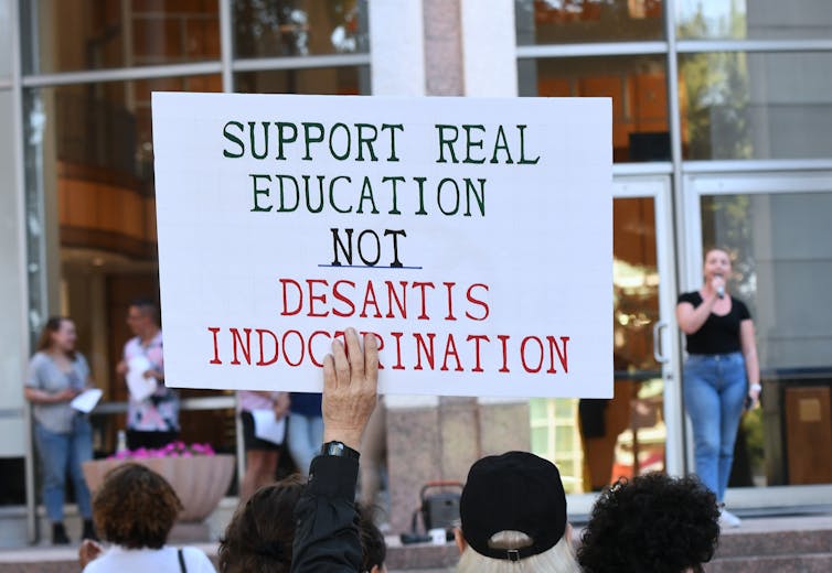 A woman speaking into a microphone stands on a brick ledge. People holding a sign that reads: 'Support Real Education Not DeSantis Disinformation' face her from the ground below.