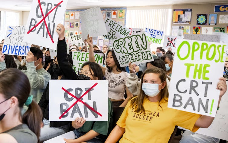 Teenage girls and boys, some wearing face masks, stand together, holding signs opposing bans on critical race theory.