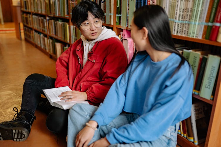 Two students sit, talking in a library.