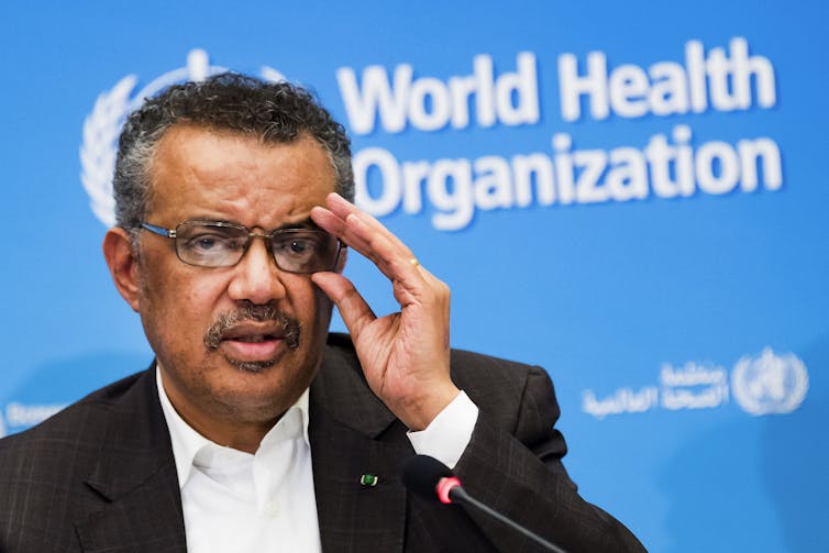 A man with a moustache adjusts his glasses in front of a World Health Organization logo on a blue wall.