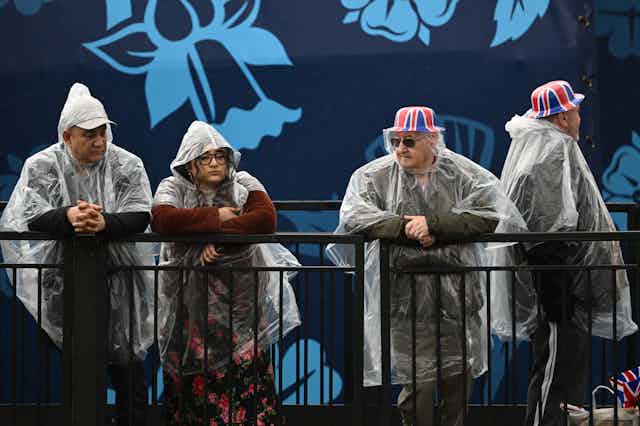 Four people in transparent ponchos rest against a railing.