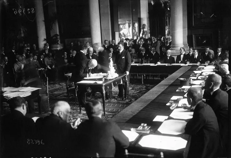 Una foto en blanco y negro que muestra a un hombre firmando un papel en una mesa rodeado de otros hombres que le observan.