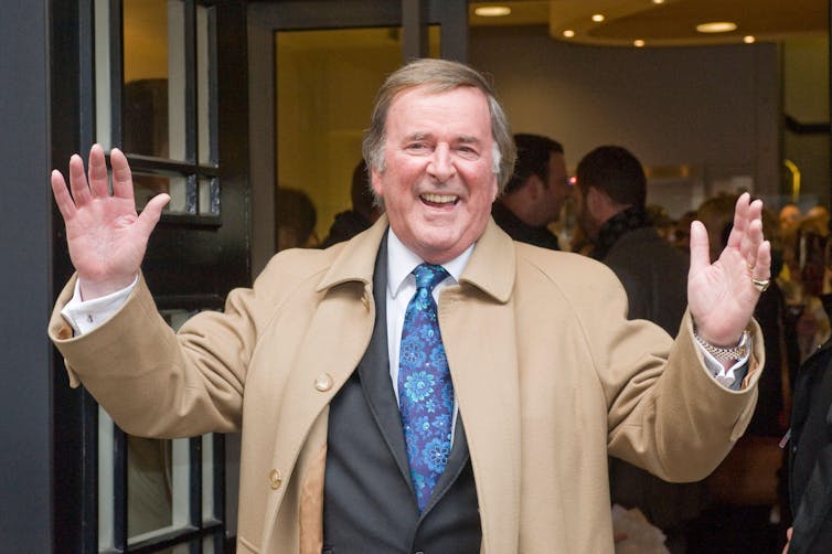 Terry Wogan holding up his hands in a beige coat.