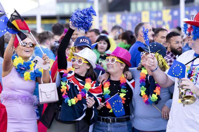 Eurovision fans wave Germany and UK flags, wearing silly glasses and hats. 