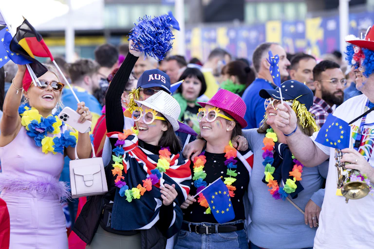 Eurovision fans wave Germany and UK flags, wearing silly glasses and hats.