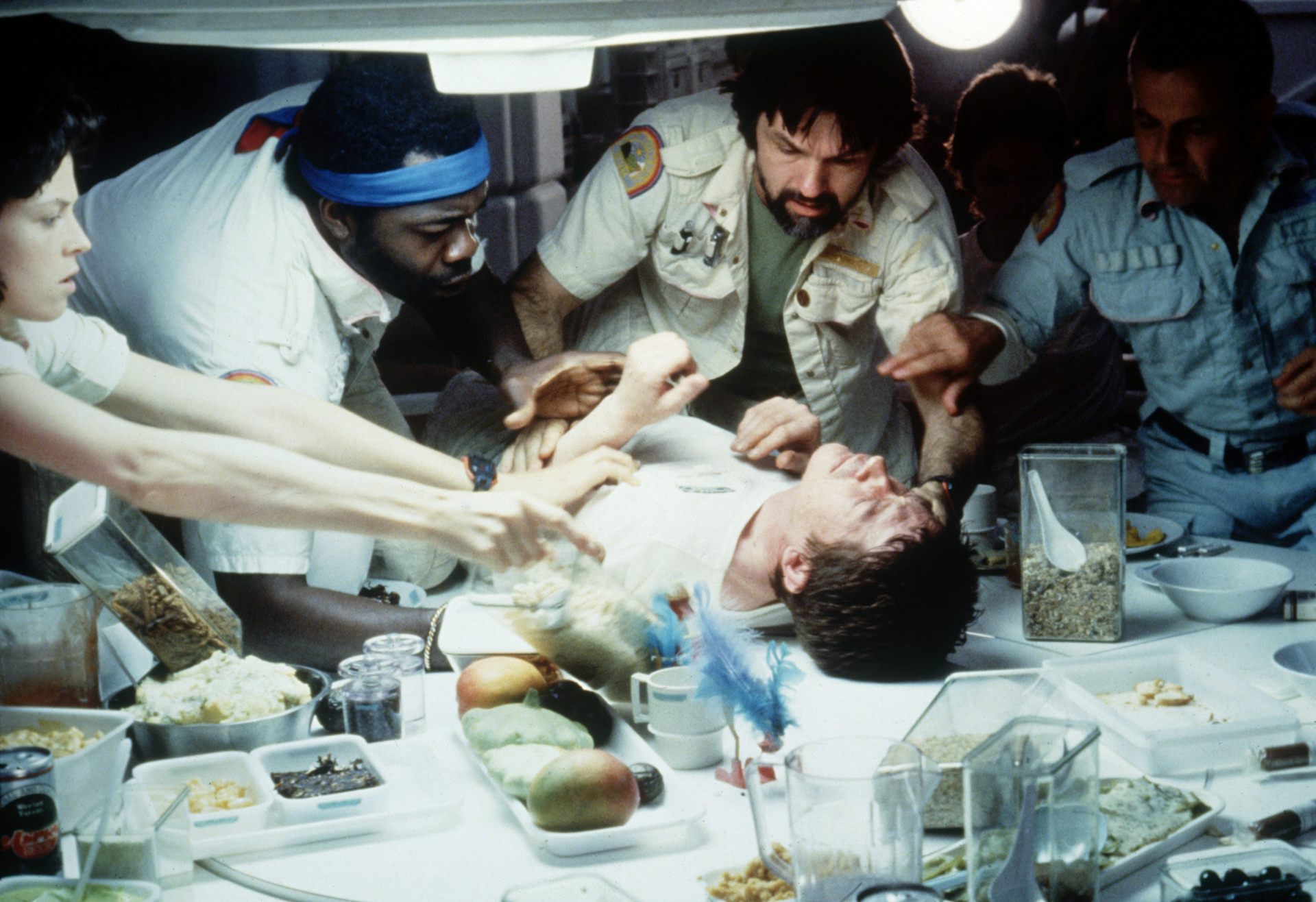 A man lies on a white table surrounded by food while four other people reach out to hold him down.