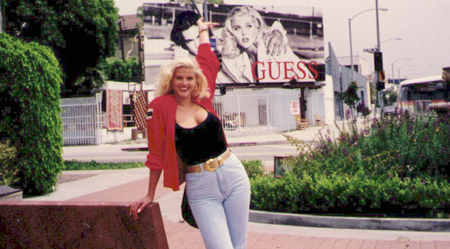 Anna Nicole Smith posing with her Guess billboard in the background. She has platinum blonde hair and wears a red shirt and blue jeans.