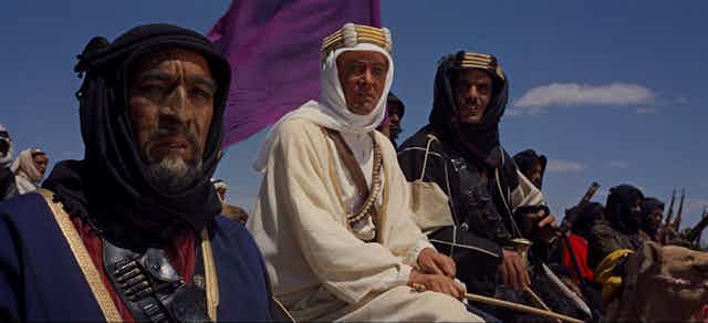 Un hombre blanco vestido con casaca y turbante a caballo junto a dos hombres árabes.