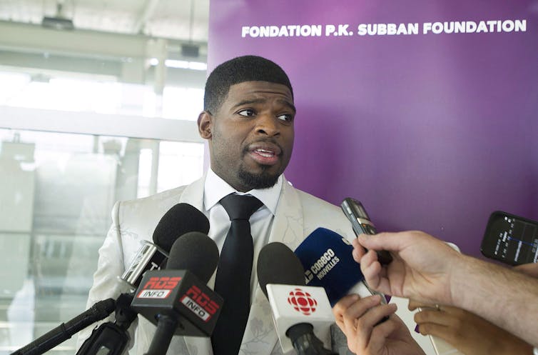 A Black man in a suit speaks to off-screen reporters in front of a bunch of hands holding microphones