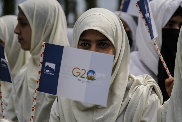 A young woman wearing a white hijab waving a small banner with the G20 logo on it.