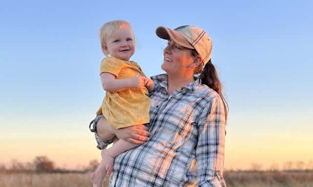 Kerissa Payne holds one of her young daughters, both smiling widely at each other, with farm fields in the background.