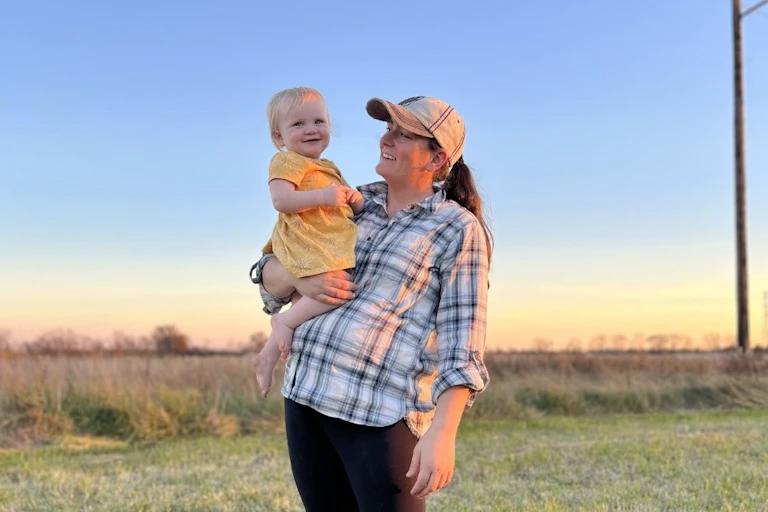 Kerissa Payne holds one of her young daughters, both smiling widely at each other, with farm fields in the background.