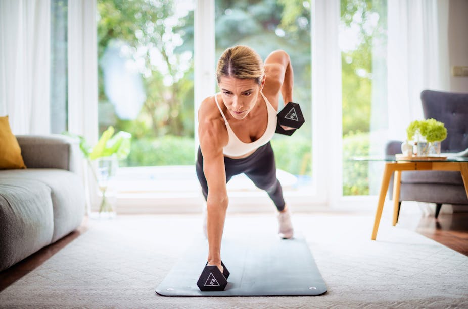 A middle-aged woman performs a plank row with dumbbells in her living room.