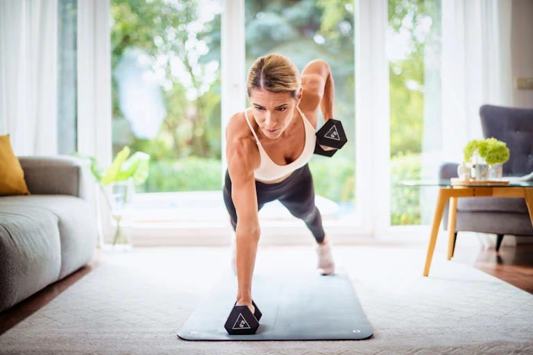 A middle-aged woman performs a plank row with dumbbells in her living room.
