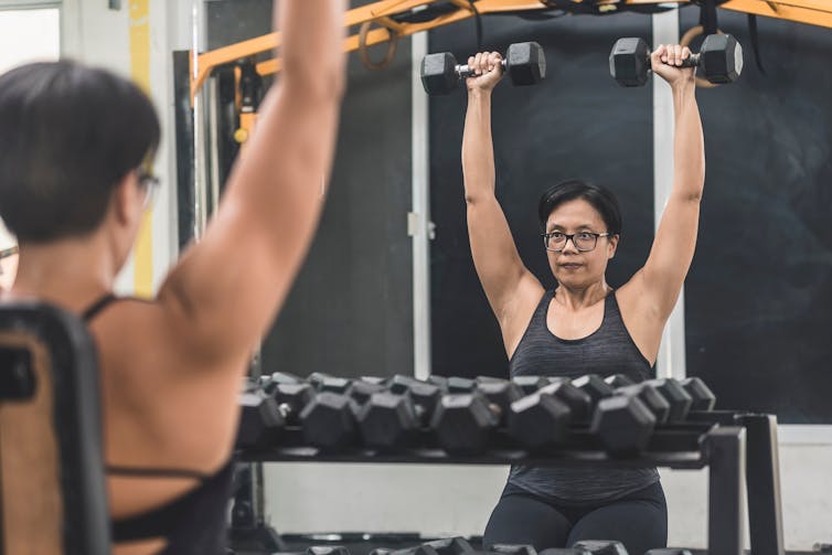 Middle aged woman performs dumbbell shoulder press in the gym.