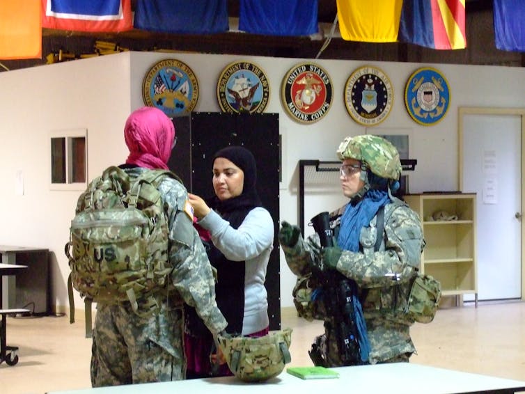 A female Afghan role-player wraps a headscarf around a female soldier while a third female soldier looks on.
