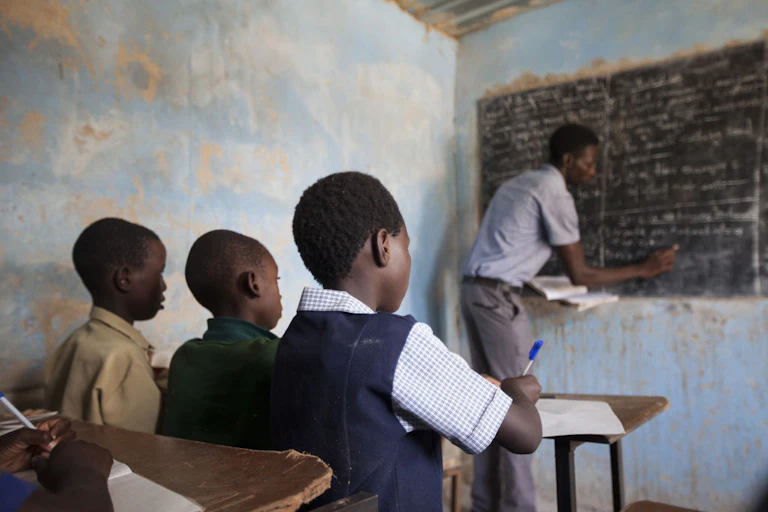 Three children sit at a school desk making notes while a teacher draws on a board with chalk at the head of the class