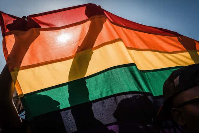 A rainbow flag is held up by several people, the sun shining through it to reveal only their silhouettes through the flag.