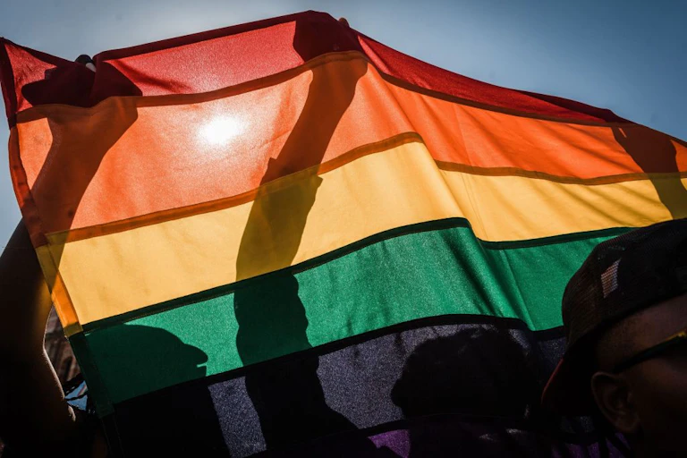 A rainbow flag is held up by several people, the sun shining through it to reveal only their silhouettes through the flag.
