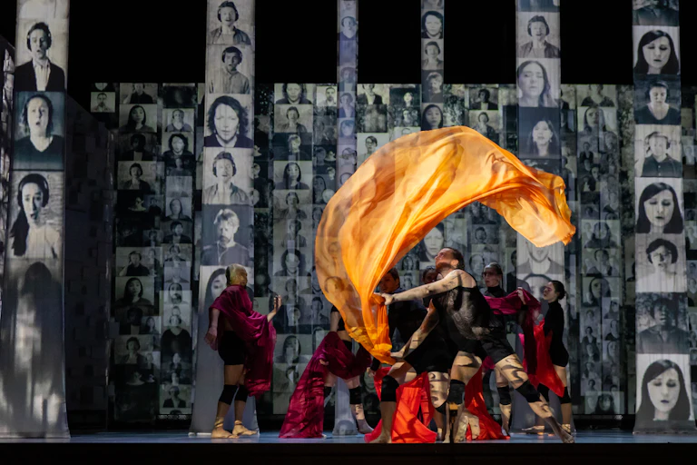 A figure on a stage seen waving a large orange silk-like fluttering sheet in the air, surrounded by black and white portraits in rows of different sizes