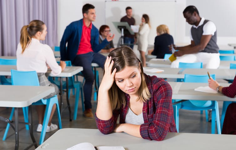 A student seen sitting at a desk holding their head while people are talking behind them.