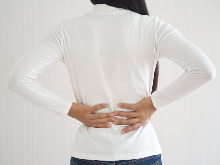 A person wearing a long sleeved white top faces away from the camera clasping the bottom of their back.